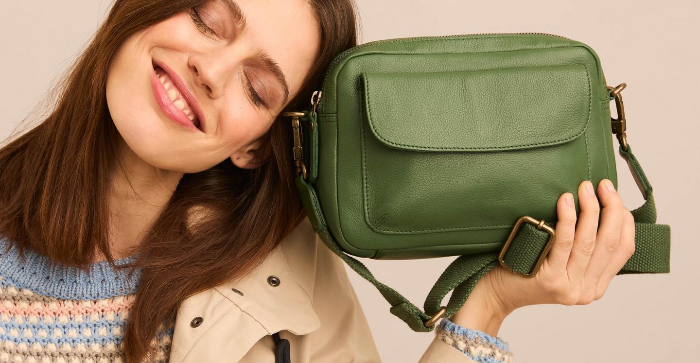 A woman holding up a green leather camera bag with zip top,large front pocket & adjustable webbed canvas strap.