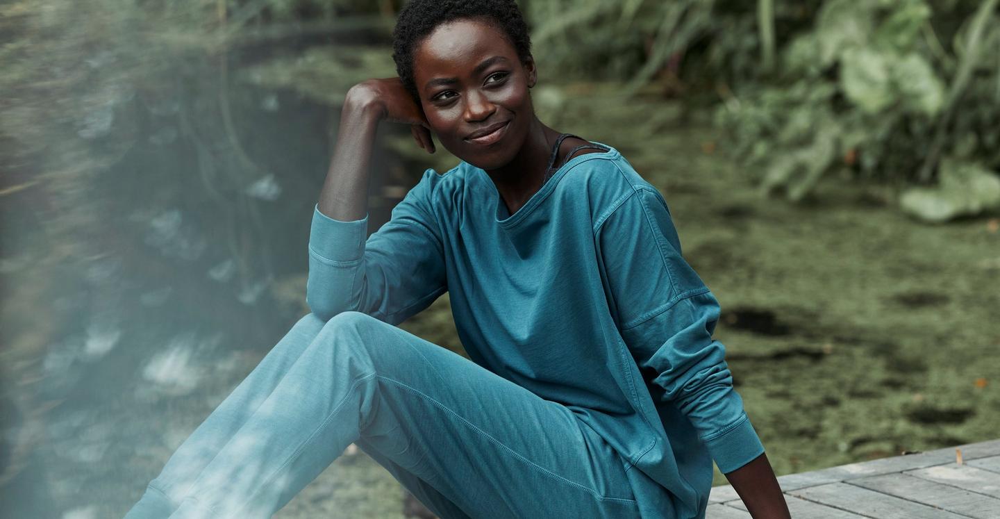 A woman sitting in a green garden, wearing a blue relaxed-fit top & co-ordinating joggers.
