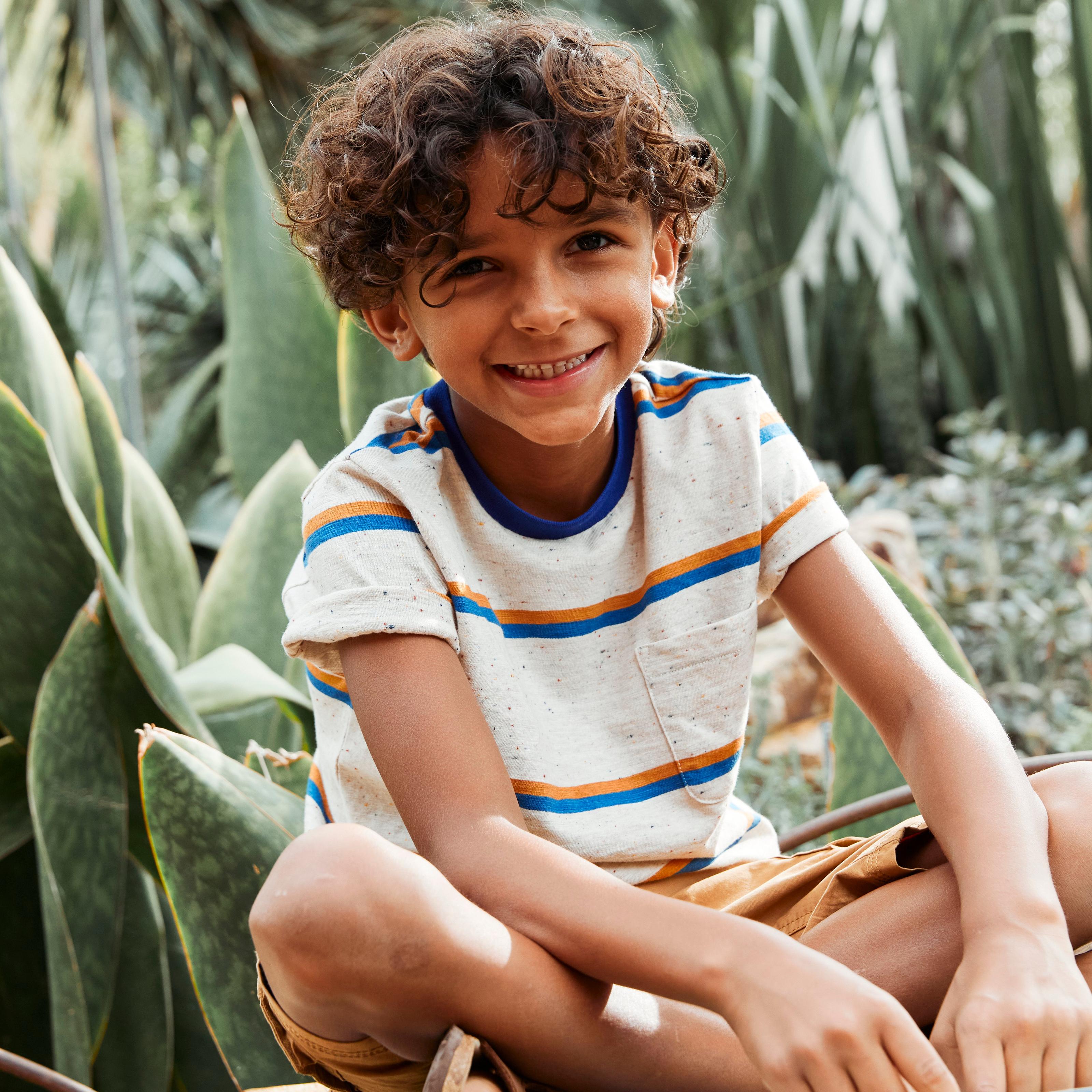 A boy wearing a flecked ivory T-shirt with blue & yellow stripes & chest pocket, & mustard-yellow shorts.