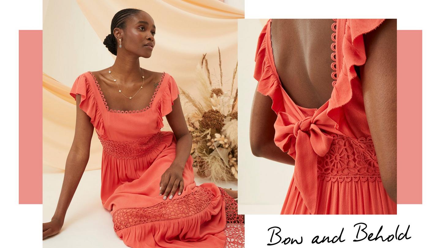 A woman wearing a coral-pink maxi dress with a square neckline, flutter sleeves, floral lace panels & tied bow on the back.