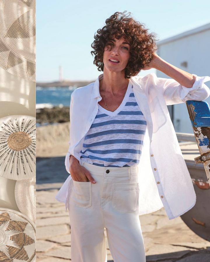 A woman wearing an oversized white linen shirt, white & blue striped V-neck T-shirt, & high-waisted ivory coloured trousers.