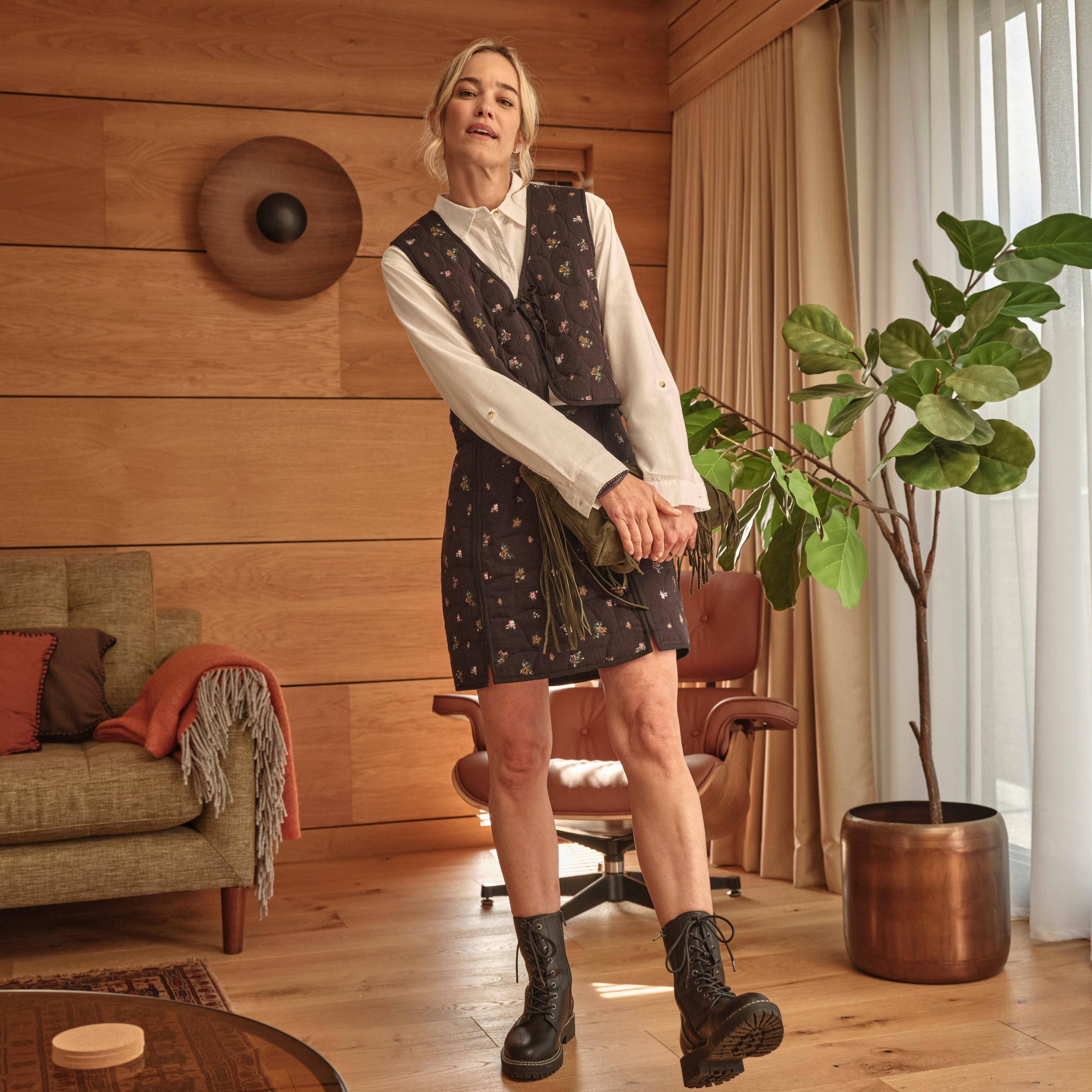 A woman wearing a dark brown padded waistcoat & skirt with floral print, a long-sleeved white shirt & black leather boots.