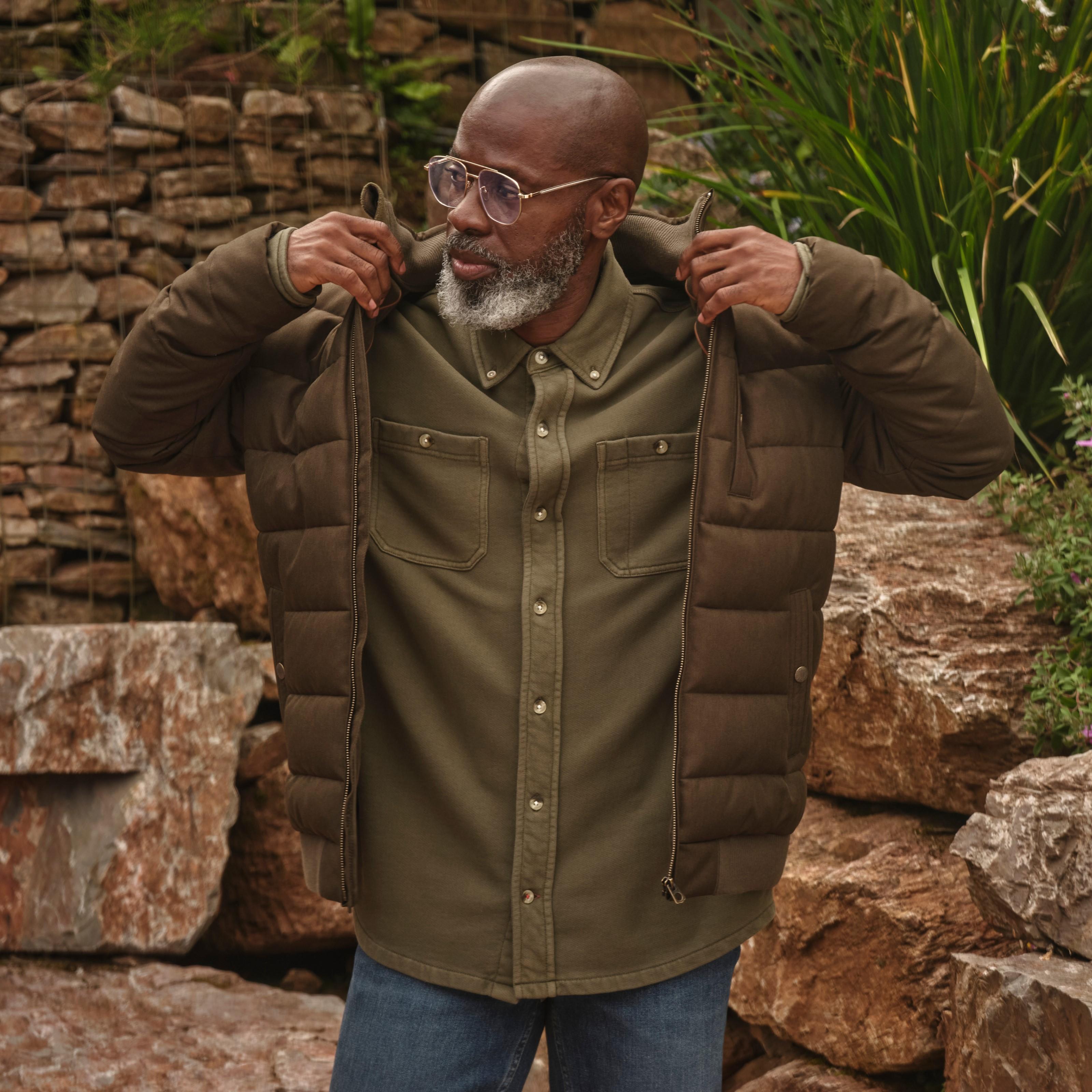 A man wearing a brown padded jacket over a khaki-green long-sleeved shirt & blue jeans.