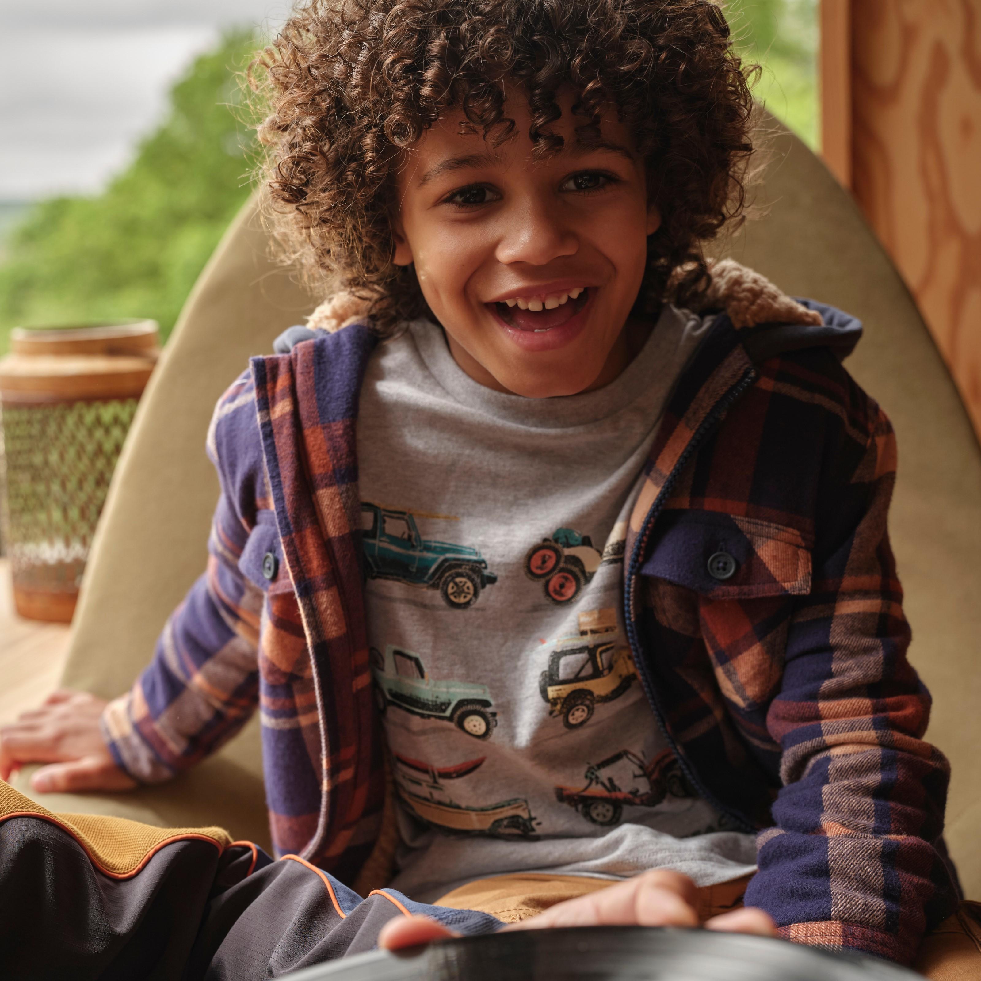 A boy wearing a navy blue & orange checked shirt over a Land Rover graphic T-shirt & tan brown chinos.