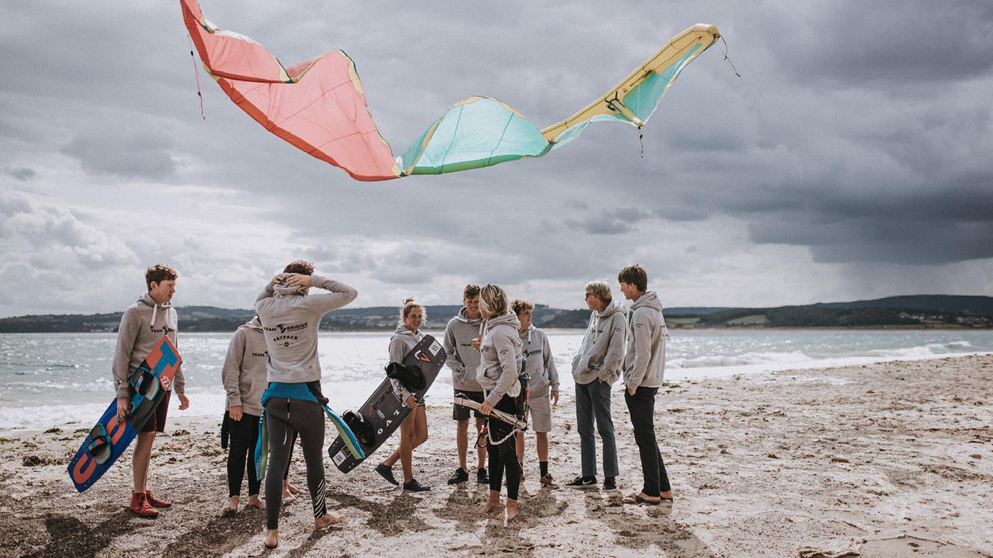A kite floats in the air above the kitesurfers stood on a sandy beach
