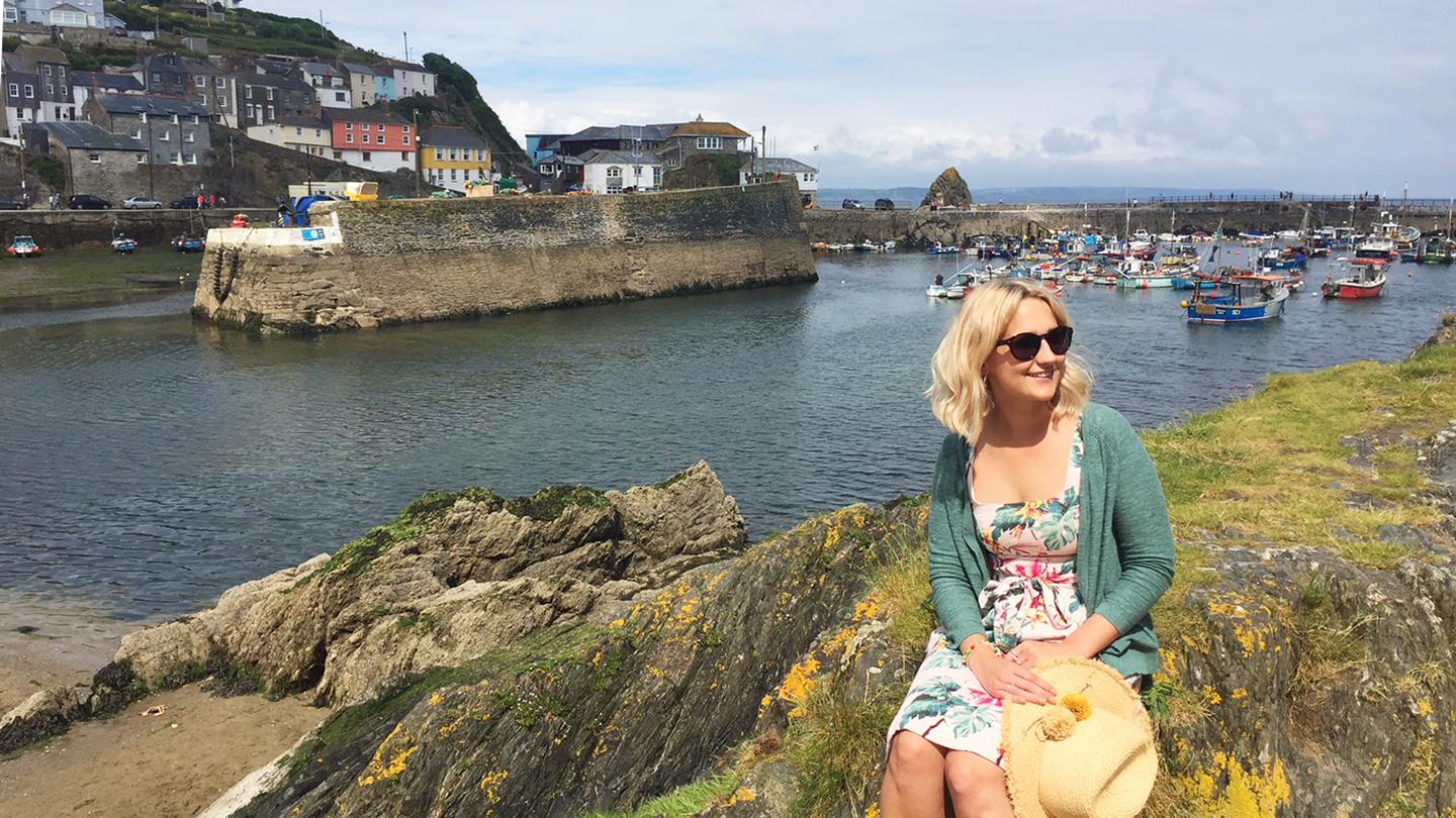 Grace, store manager at Mevagissey FatFace, sitting by the beautiful Cornish harbour wearing FatFace clothes