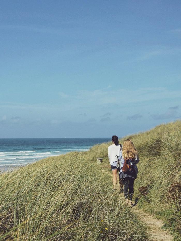 Two women walking along a grassy coastal path, beneath a blue sky.