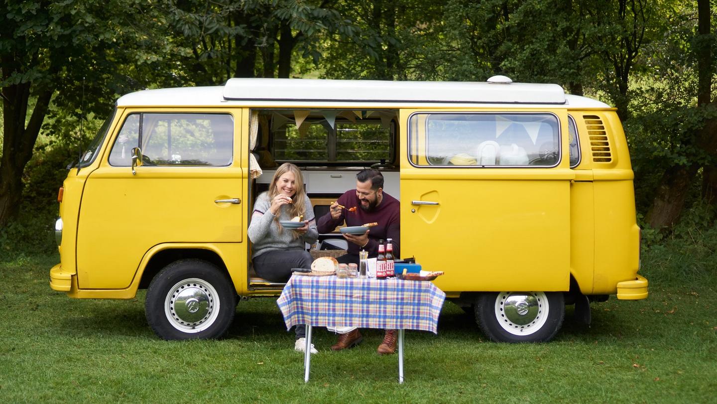 Blonde girl wearing a grey jumper and dark haired male wearing a red jumper sat on the edge of a yellow campervan eating food from bowls
