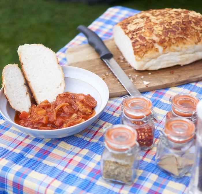 A table with a bowl of pork and chorizo stew, a loaf of bread on a board and 4 small pots of herbs and spices on it