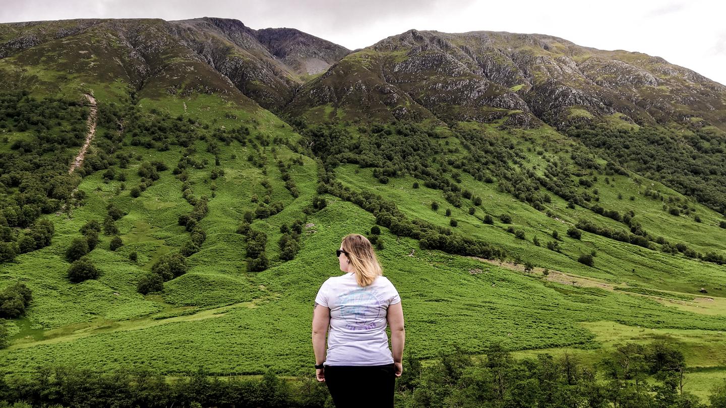 Sarah from the FatFace store in Fort William, wearing a FatFace t-shirt while standing in front of beautiful, lush green rolling hills